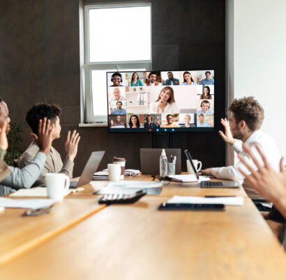 Diverse businesspeople making online video call to employees Distant Virtual Meeting Concept. Diverse business people making online video call with colleagues, sitting at desk in boardroom, waving hands at tv screen at office, talking on web with remote workers